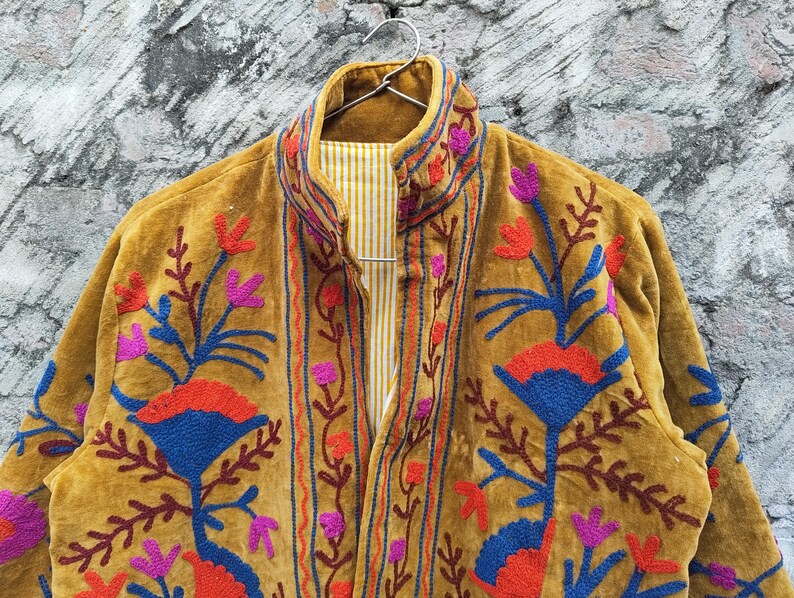 May include: Mustard-coloured jacket with detailed floral embroidery in red, blue, and purple. The jacket has a high collar and a striped lining. The embroidery provides a striking contrast against the jacket's textured fabric, showcasing a bohemian style.
