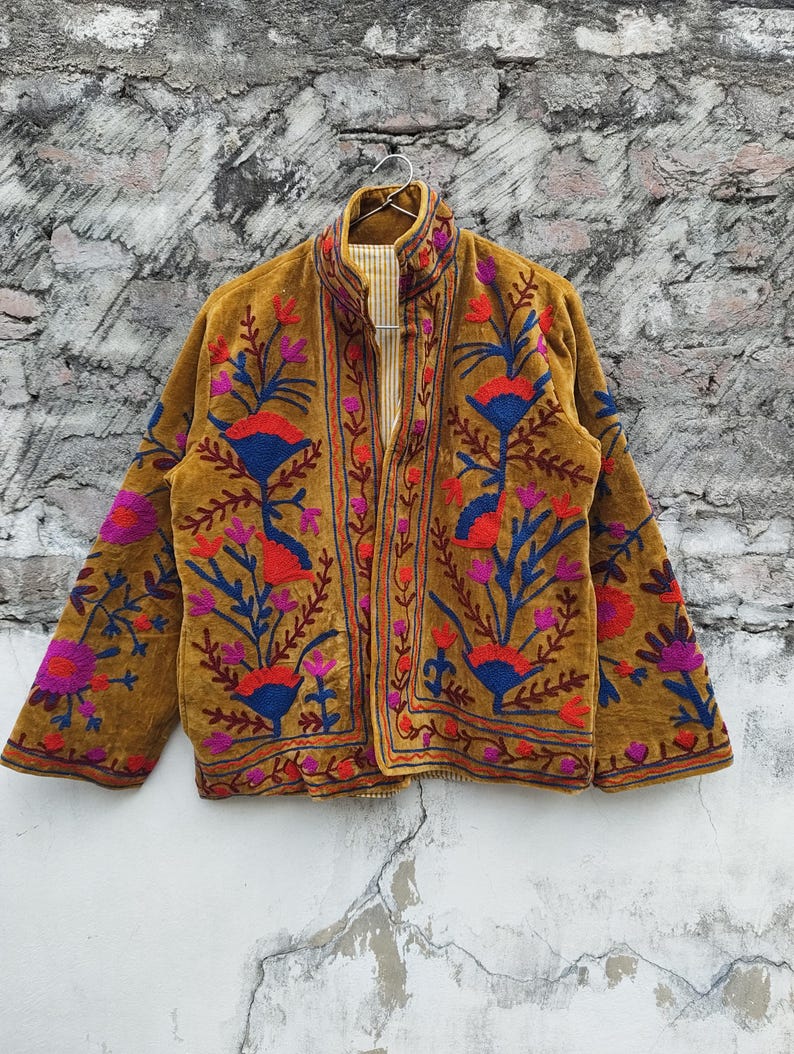 May include: Mustard-coloured jacket with detailed floral embroidery in red, blue, and purple. The jacket has a high collar and a striped lining. The embroidery forms a detailed pattern across the front and sleeves.