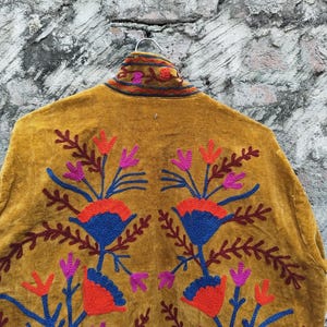 May include: Mustard-coloured jacket featuring detailed floral embroidery in red, blue, and purple. The jacket hangs on a metal hanger against a textured grey background. The embroidery is symmetrical, with floral patterns on the back.