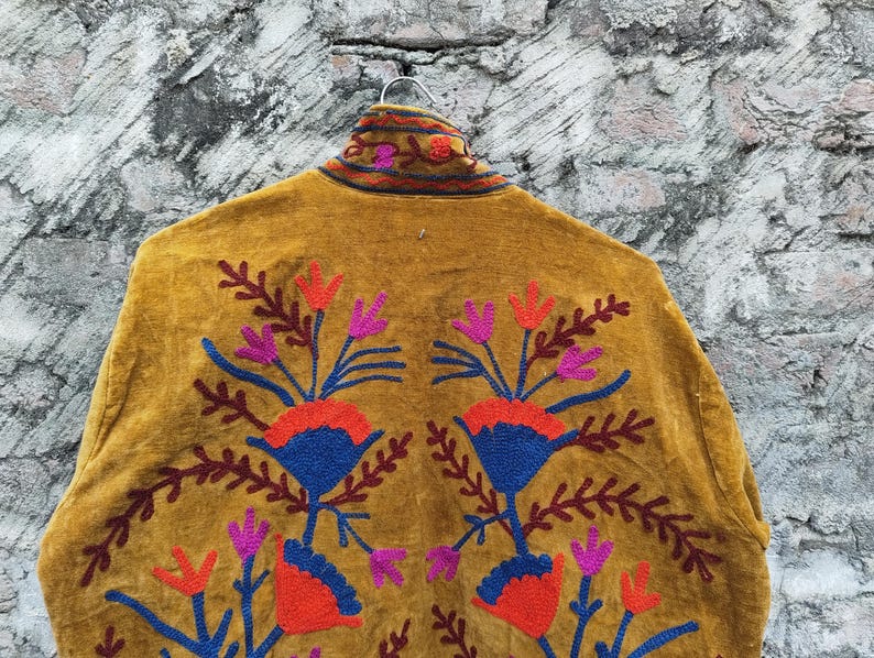May include: Mustard-coloured jacket featuring detailed floral embroidery in red, blue, and purple. The jacket hangs on a metal hanger against a textured grey background. The embroidery is symmetrical, with floral patterns on the back.
