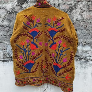 May include: Mustard-coloured jacket with detailed floral embroidery in red, blue, and purple. The jacket has a high collar and patterns along the hem and back, creating a bohemian aesthetic.
