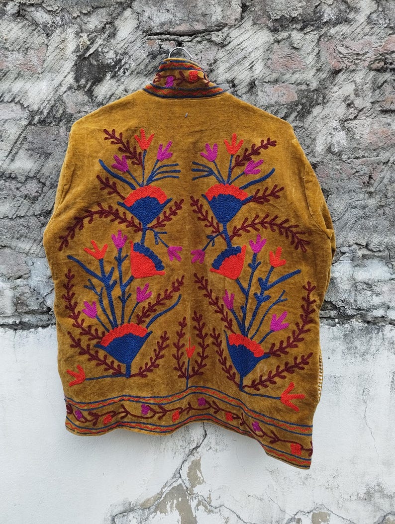 May include: Mustard-coloured jacket with detailed floral embroidery in red, blue, and purple. The jacket has a high collar and patterns along the hem and back, creating a bohemian aesthetic.