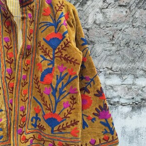 May include: Mustard-coloured jacket with detailed floral embroidery in red, blue, and purple. The jacket has a high collar and a striped lining. The embroidery covers the front and sleeves. The jacket is a vibrant piece of clothing.