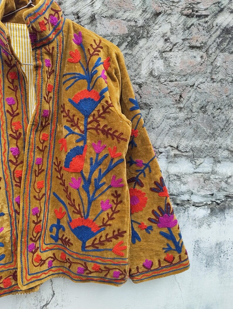 May include: Mustard-coloured jacket with detailed floral embroidery in red, blue, and purple. The jacket has a high collar and a striped lining. The embroidery covers the front and sleeves. The jacket is a vibrant piece of clothing.