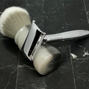 May include: A silver safety razor with a white and gray handle and a matching brush with a white bristle head. The razor and brush are on a black marble surface.