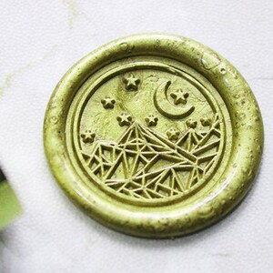 May include: A gold wax seal featuring a design of a crescent moon, stars, and geometric mountains.