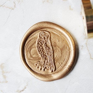 Puede incluir: Un sello de cera dorado con un diseño de búho. El sello es redondo y tiene un diseño de búho en relieve en el centro. El búho está de frente y tiene las alas extendidas. El sello está sobre un fondo blanco.