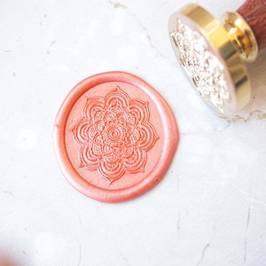 May include: A coral-coloured wax seal featuring a detailed mandala design. A gold-coloured metal stamp with a wooden handle is positioned nearby. The seal is on a white surface, suggesting stationery or craft supplies.