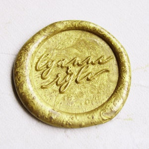 Könnte beinhalten: Ein goldenes Siegelwachs mit der Inschrift "Lyann & Kyle" in Kursivschrift.