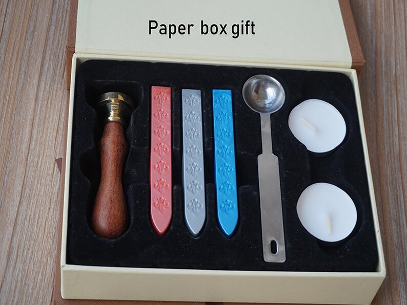 Peut inclure: Un coffret cadeau en papier avec un tampon &agrave; cacheter &agrave; manche en bois, trois b&acirc;tons de cire color&eacute;s, une cuill&egrave;re &agrave; fondre en m&eacute;tal et deux bougies chauffe-plat blanches. La bo&icirc;te est beige et porte l'inscription "Paper box gift".