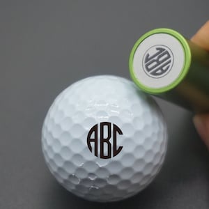 May include: A white golf ball with a black monogram "ABC" printed on it. A green golf ball stamp with a monogram "JDB" is shown in the background.