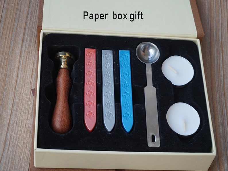 May include: A set of wax sealing supplies in a white box with a black interior. The set includes a wooden handle with a brass seal head, four wax sticks in red, gray, blue, and silver, a metal spoon, and two white tealights.