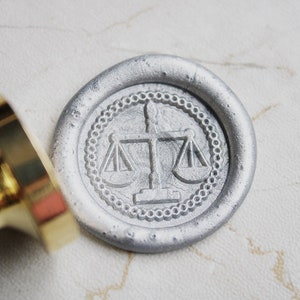 Puede incluir: Un sello de cera plateado con un diseño en relieve de una balanza de la justicia. El sello está sobre una superficie blanca.