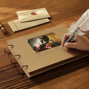May include: A brown leather-bound photo album with metal rings. The album is open to a page with a photo of a flower and a handwritten message in Japanese. A white feather pen is resting on the page.