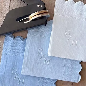 May include: A black metal embosser tool positioned above three paper napkins with scalloped edges. Two napkins are light blue and one is white. Each napkin features an embossed monogram design with the letters S and N separated by an ampersand.