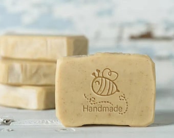May include: A stack of four rectangular bars of soap with a bee design and the word "Handmade" on the top bar. The soap is a light beige color.