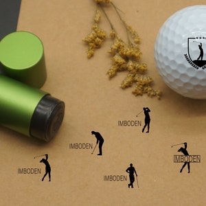 May include: A green golf ball stamper with the word "IMBODEN" printed on the side. The stamper is used to mark golf balls with the name "IMBODEN".