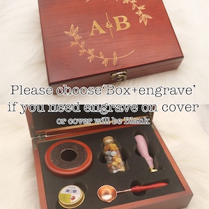 Puede incluir: Una caja de madera con tapa con bisagras, grabada con las letras "A&B" en fuente dorada. La caja est&aacute; abierta, revelando un juego de suministros para sellos de cera, que incluyen una cuchara para derretir cera, un frasco de cuentas de cera, un sello de cera y un peque&ntilde;o frasco de cera para sellar.