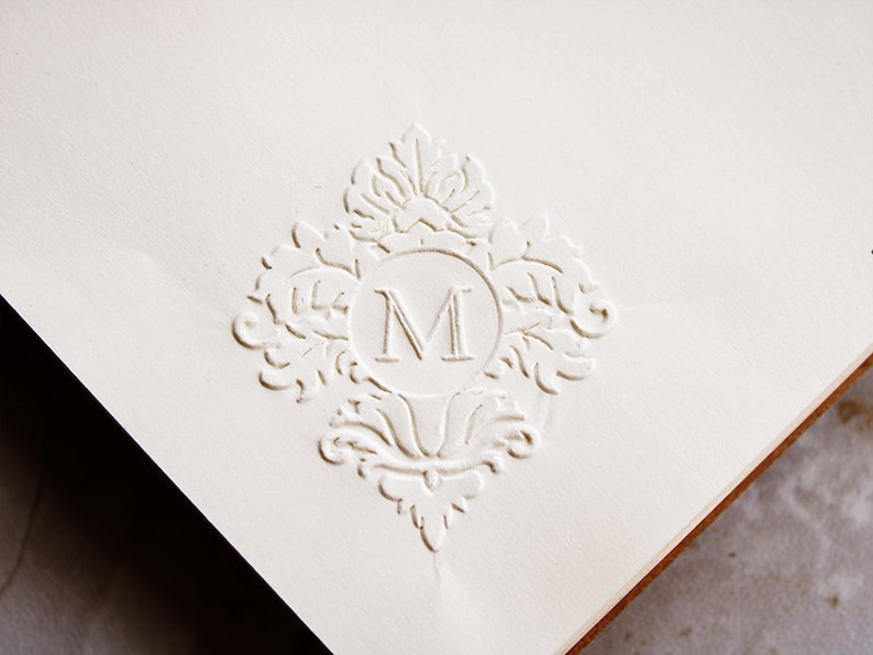 May include: A white embossed paper with a decorative design featuring a large letter "M" in the center.
