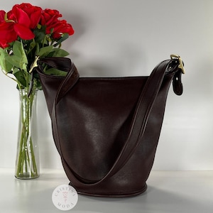 May include: A dark burgundy leather bucket bag with a shoulder strap and gold-tone hardware. The bag is empty and stands upright. A bouquet of red roses in a clear glass vase is to the left.