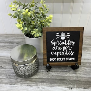 May include: A wooden sign with a black background and white text that reads "Sprinkles are for cupcakes (not toilet seats)". The sign has a small cupcake graphic with white frosting and a cherry on top. The sign is sitting on a wooden table with a small potted plant and a silver jar.