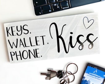 May include: White wooden sign with black text that reads "Keys. Wallet. Phone. Kiss" with a small blue heart above the word "Kiss".