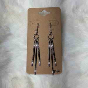 May include: A pair of dangle earrings with black beads and silver-tone metal accents. The earrings have a vertical design with multiple strands of black beads, interspersed with white and purple beads. They are displayed on a brown card.