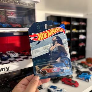 May include: A Hot Wheels die-cast model car in a blister pack. The car is primarily orange and white with the number 56 on the side. The packaging features the Hot Wheels logo and an image of a woman in a bikini. Other toy cars are visible in the background.