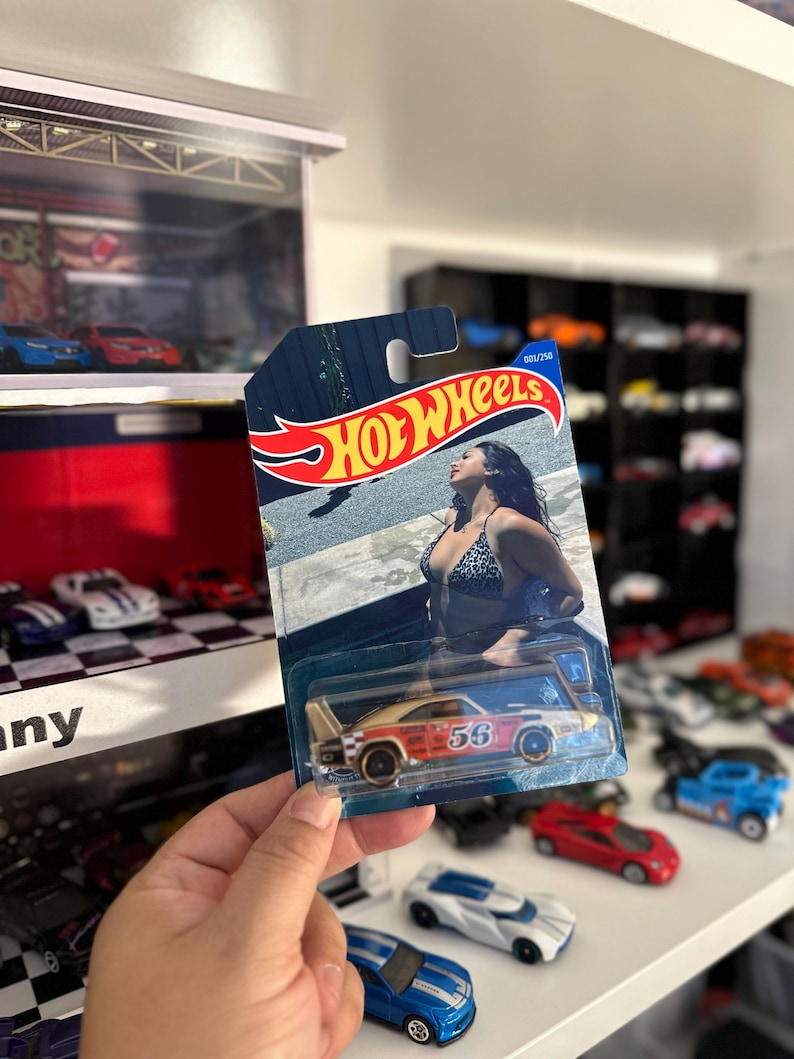 May include: A Hot Wheels die-cast model car in a blister pack. The car is primarily orange and white with the number 56 on the side. The packaging features the Hot Wheels logo and an image of a woman in a bikini. Other toy cars are visible in the background.