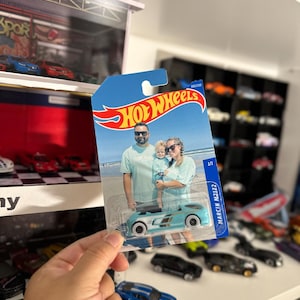 May include: A Hot Wheels die-cast car in a blue and white color scheme, held in a person's hand. The packaging features a family photo and the Hot Wheels logo. Other toy cars are visible in the background.