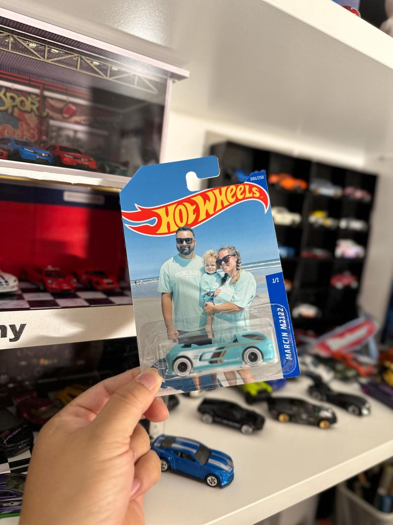 May include: A Hot Wheels die-cast car in a blue and white color scheme, held in a person's hand. The packaging features a family photo and the Hot Wheels logo. Other toy cars are visible in the background.