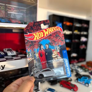 May include: A Hot Wheels die-cast toy car in a package. The package features a red and gold design with the Hot Wheels logo and an image of a couple. The car is silver with black wheels. The package reads "ALEXANDRE".