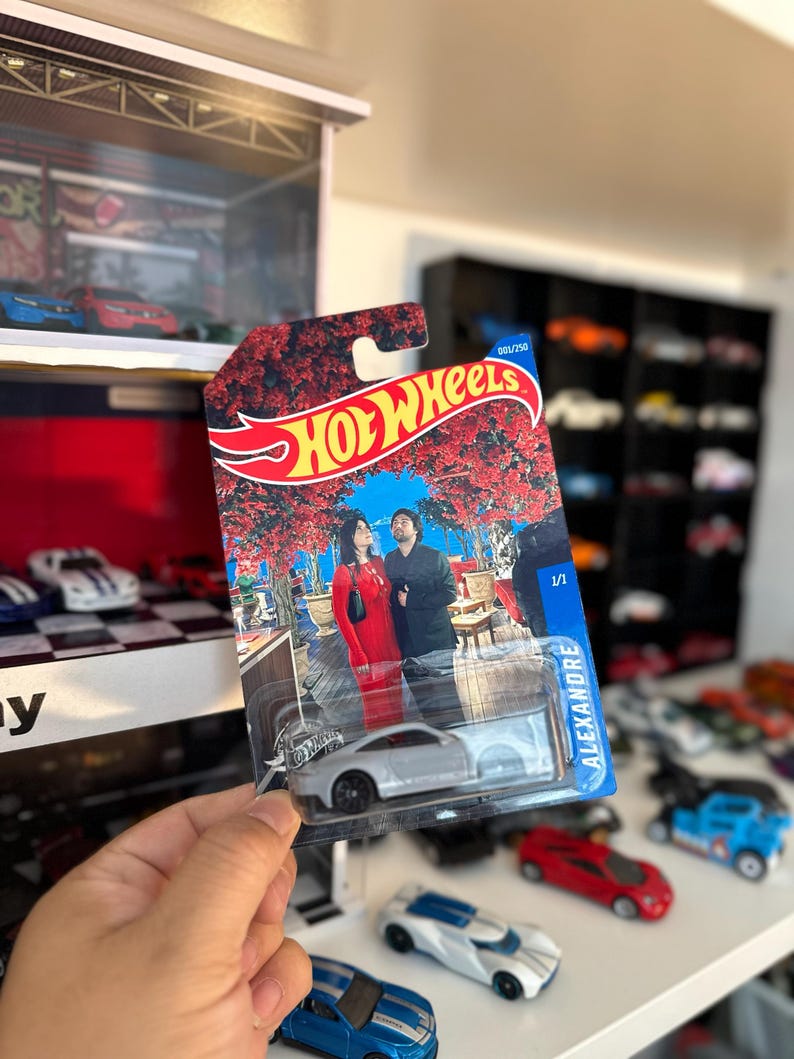 May include: A Hot Wheels die-cast toy car in a package. The package features a red and gold design with the Hot Wheels logo and an image of a couple. The car is silver with black wheels. The package reads "ALEXANDRE".