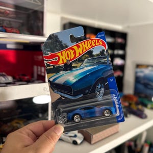 May include: A blue Hot Wheels die-cast model car in its original packaging. The packaging features a picture of a blue and white classic car. The car is a 1/1 scale model.