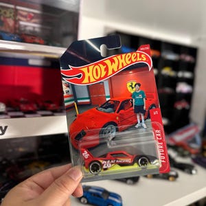 May include: A Hot Wheels die-cast toy car in a blister pack. The packaging features a red Ferrari and the Hot Wheels logo. The toy car is red, black, and yellow with the number 26. The words "FUTURE CAR" are printed on the side.