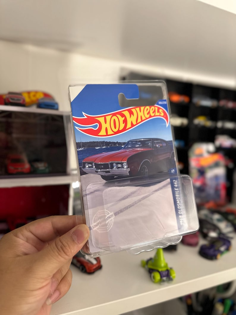 May include: A Hot Wheels die-cast car package featuring a red 1968 Oldsmobile 442. The package has a blue and white background with the Hot Wheels logo. The car is visible through the clear plastic.