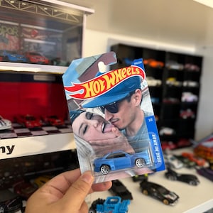 May include: A blue Hot Wheels die-cast car in its original packaging. The packaging features a photo of a couple and the text "XOXO MI AMOR". The car is a coupe with silver wheels.