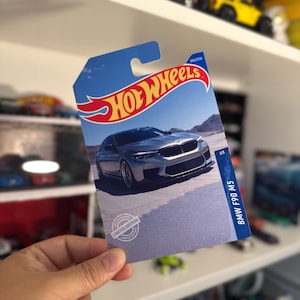 May include: A Hot Wheels card featuring a silver BMW F90 M5 car on a desert landscape. The card has a blue background with the Hot Wheels logo in red and yellow. The card is labeled "Limited Edition."