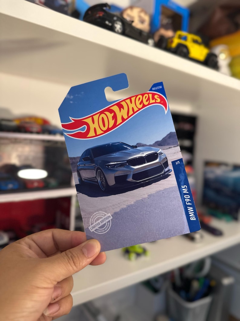 May include: A Hot Wheels card featuring a silver BMW F90 M5 car on a desert landscape. The card has a blue background with the Hot Wheels logo in red and yellow. The card is labeled "Limited Edition."