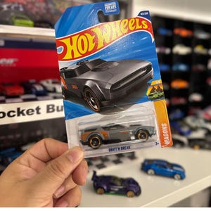 May include: A gray Hot Wheels die-cast car in its original packaging. The car is a Drift'n Break model, part of the Wagons series, and is number 183 of 250. The packaging features the Hot Wheels logo and the text "GUARANTEED FOR LIFE".
