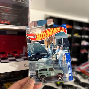 May include: A Hot Wheels die-cast model of a light green Land Rover Defender 90, with the number 68 on the side. The packaging features a photo of a lion on a car hood and a man holding a child. The text "HOT WHEELS" is visible.