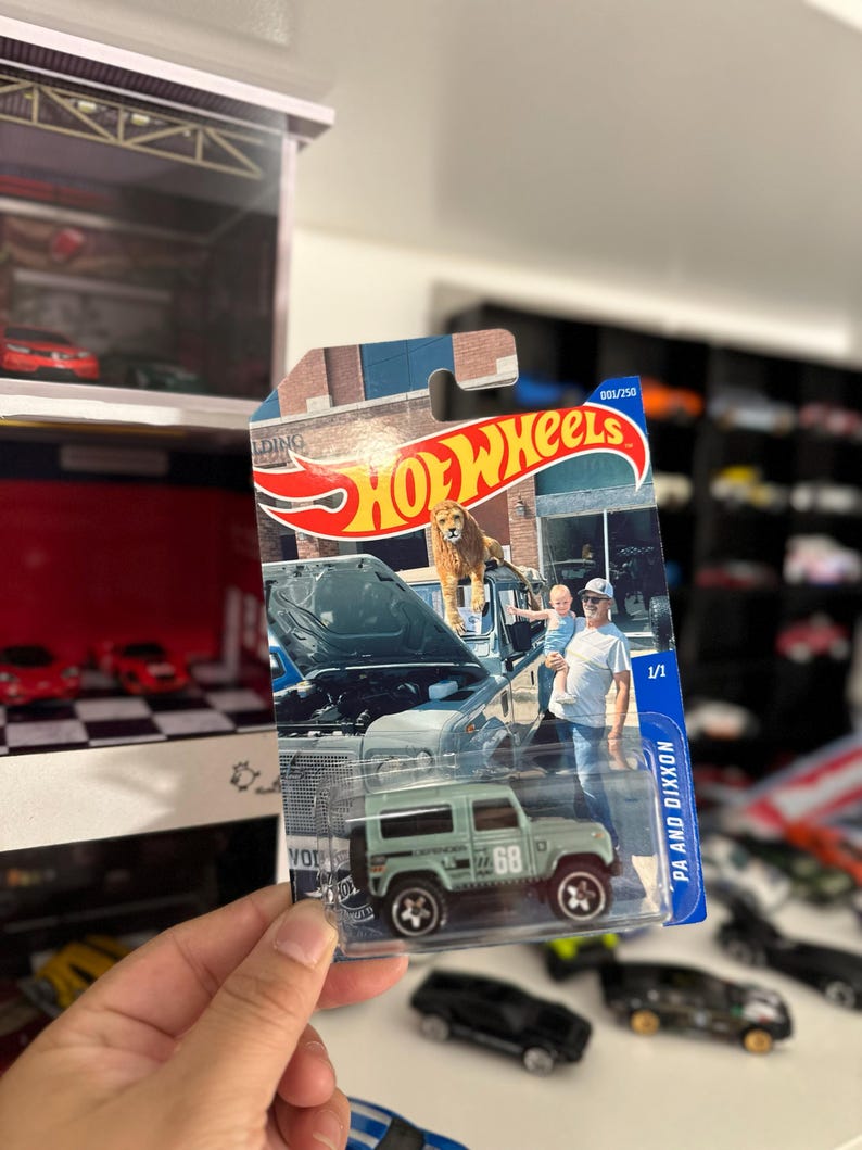 May include: A Hot Wheels die-cast model of a light green Land Rover Defender 90, with the number 68 on the side. The packaging features a photo of a lion on a car hood and a man holding a child. The text "HOT WHEELS" is visible.