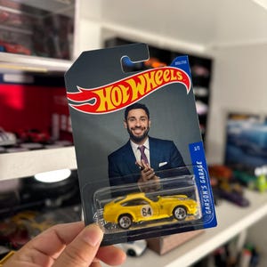 May include: A yellow Hot Wheels die-cast car with the number 64 on the side, packaged on a card featuring a man in a suit. The card has the Hot Wheels logo and the text "GARSON'S GARAGE".