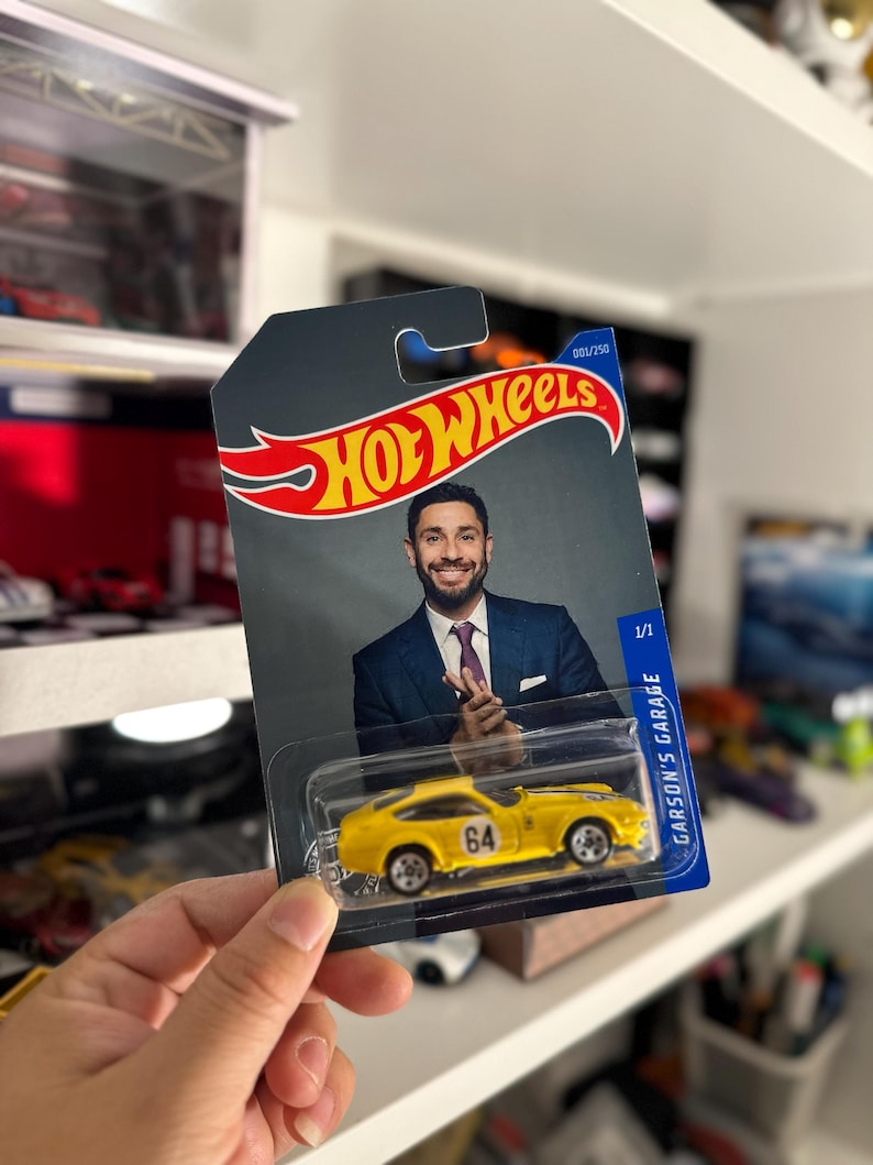 May include: A yellow Hot Wheels die-cast car with the number 64 on the side, packaged on a card featuring a man in a suit. The card has the Hot Wheels logo and the text "GARSON'S GARAGE".