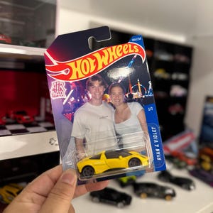 May include: A yellow Hot Wheels toy car in its original packaging. The packaging features a photo of a couple and the text "HOT WHEELS" in red and yellow. The card reads "RYAN & VIOLET".