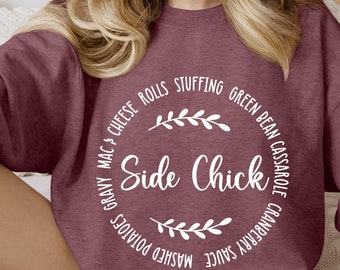 May include: A maroon sweatshirt with a white circle graphic that says "Side Chick" surrounded by the words "Gravy, Mac & Cheese Rolls, Stuffing, Green Bean Casserole, Cranberry Sauce, Mashed Potatoes".