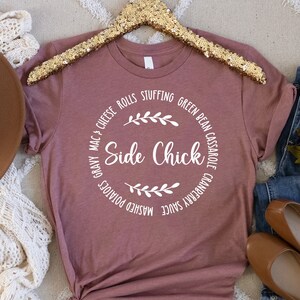 May include: A mauve t-shirt with a white circle graphic that says "Side Chick" surrounded by a list of Thanksgiving side dishes. The shirt is on a hanger and there is a brown hat, a white crocheted scarf, and blue jeans with brown flats in the background.