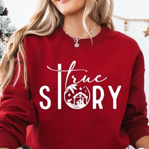 May include: A red Christmas sweatshirt with the text "True Story" in white lettering. The sweatshirt features a white illustration of a nativity scene inside a circle.