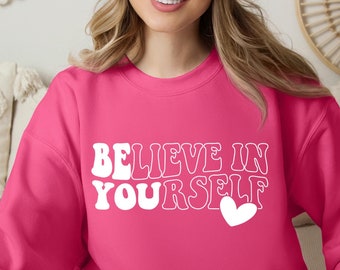 May include: A pink sweatshirt with the text "Believe in Yourself" in white outlined letters and a small white heart. The model is wearing a white beanie.