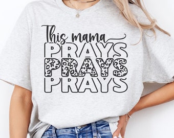 May include: A woman wearing a grey t-shirt with the text "This mama PRAYS" in black lettering with a leopard print pattern. The text is stacked vertically with the word "PRAYS" repeated three times.